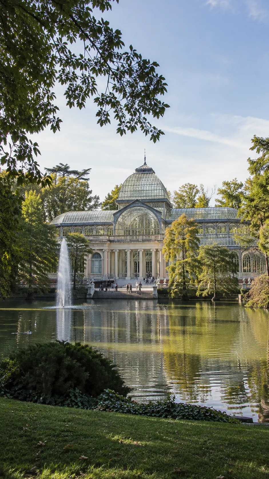 Palacio de Cristal del parque del Retiro reflejado en su estanque, con el chorro de agua y los árboles alrededor